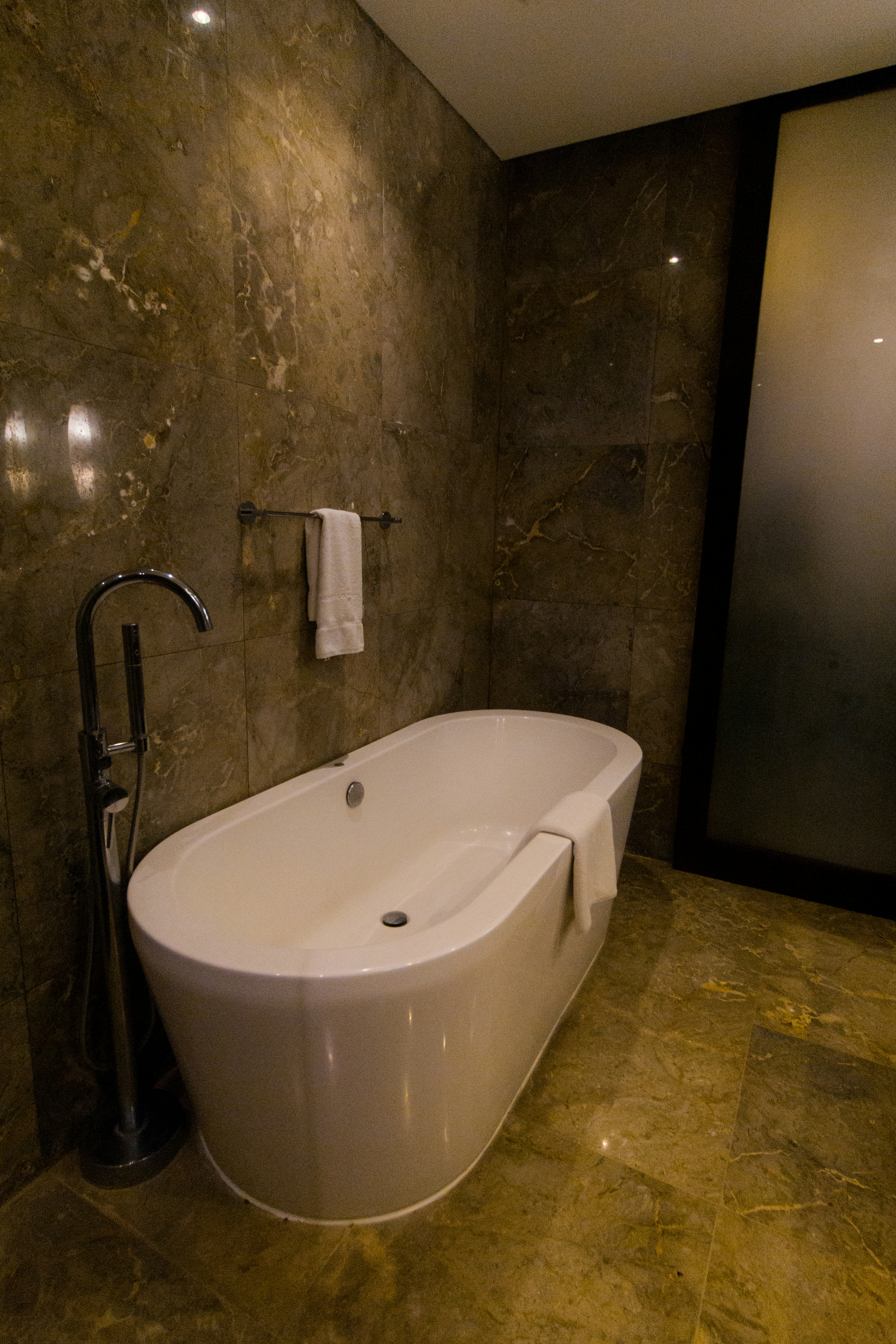 A modern bathroom featuring a sleek, freestanding bathtub with a curved design. The walls and floor have a marble finish in various shades of brown, enhancing the luxurious feel. A white towel is draped over the side of the tub, and there is a polished silver faucet nearby. The ambient lighting casts soft reflections on the marble surfaces.