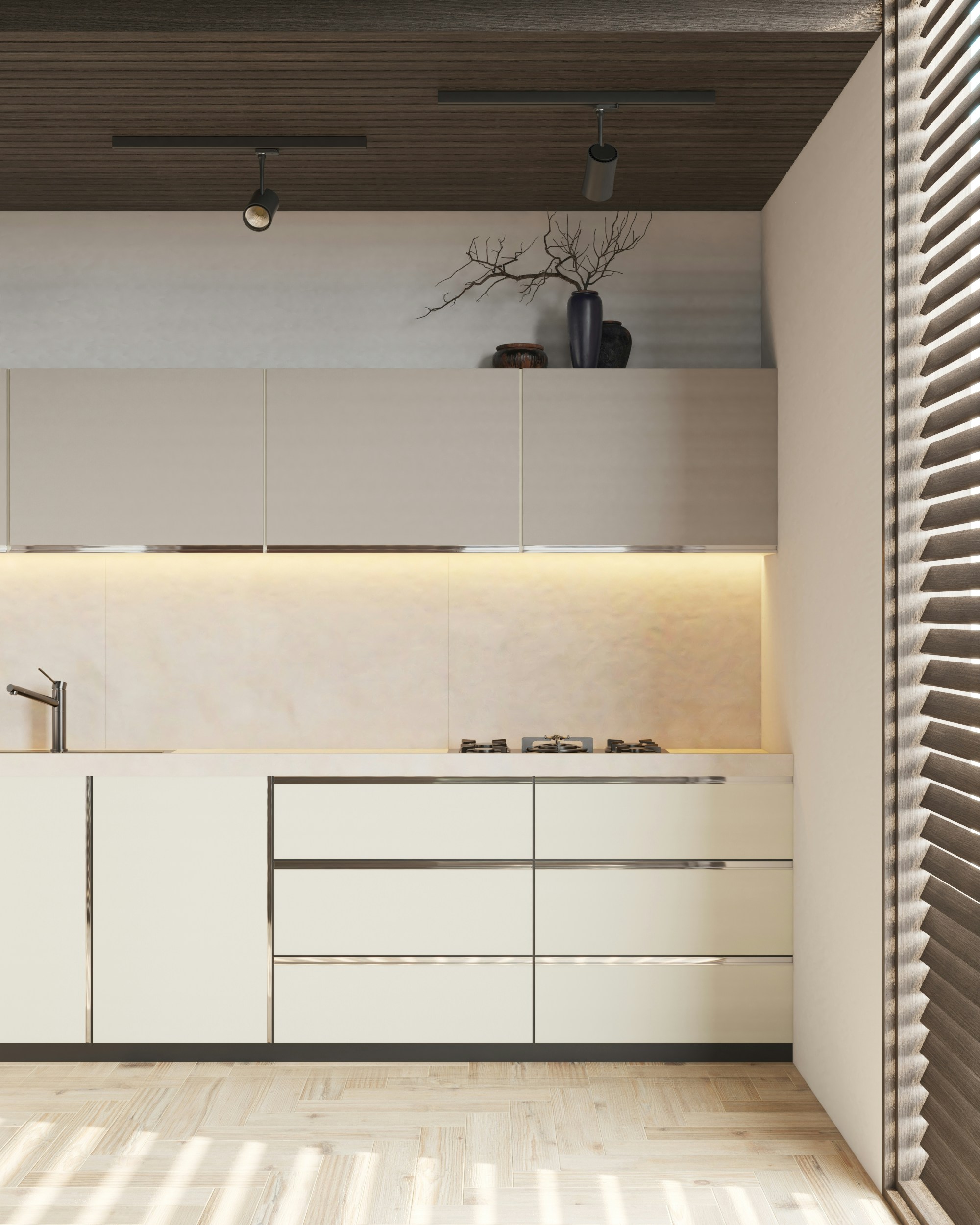 A modern kitchen with light-colored cabinets and sleek design. There are overhead cabinets, a built-in stove, and a minimalist sink. A decorative vase with branches and some pottery are placed on top of the cabinets. The ceiling features two spotlights, and there is ambient lighting under the cabinets. The floor is wooden, and the room is partially lit by sunlight coming through blinds.