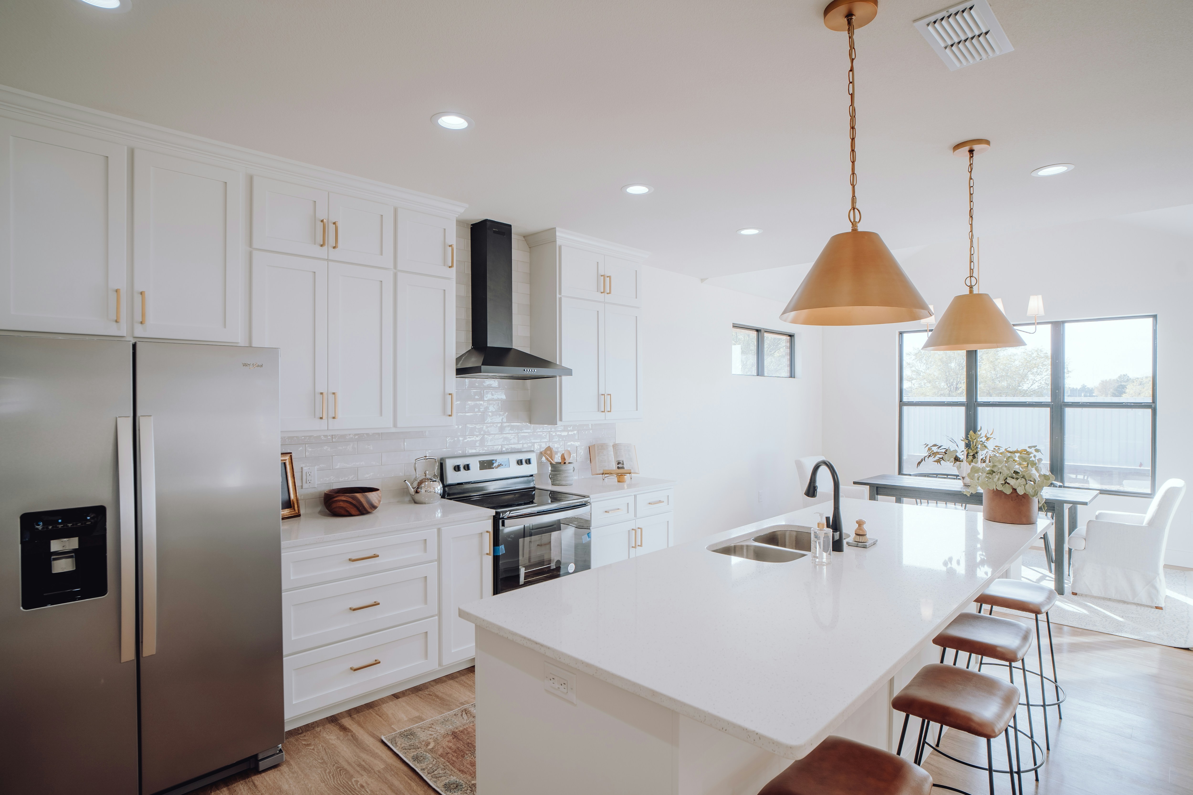A modern kitchen with white cabinetry and a large island with barstools. The room features stainless steel appliances including a refrigerator and oven. Copper pendant lights hang over the island, which has a sink and a vase of flowers. The space has wooden flooring and large windows allowing natural light to flood the area, creating a bright and airy atmosphere.