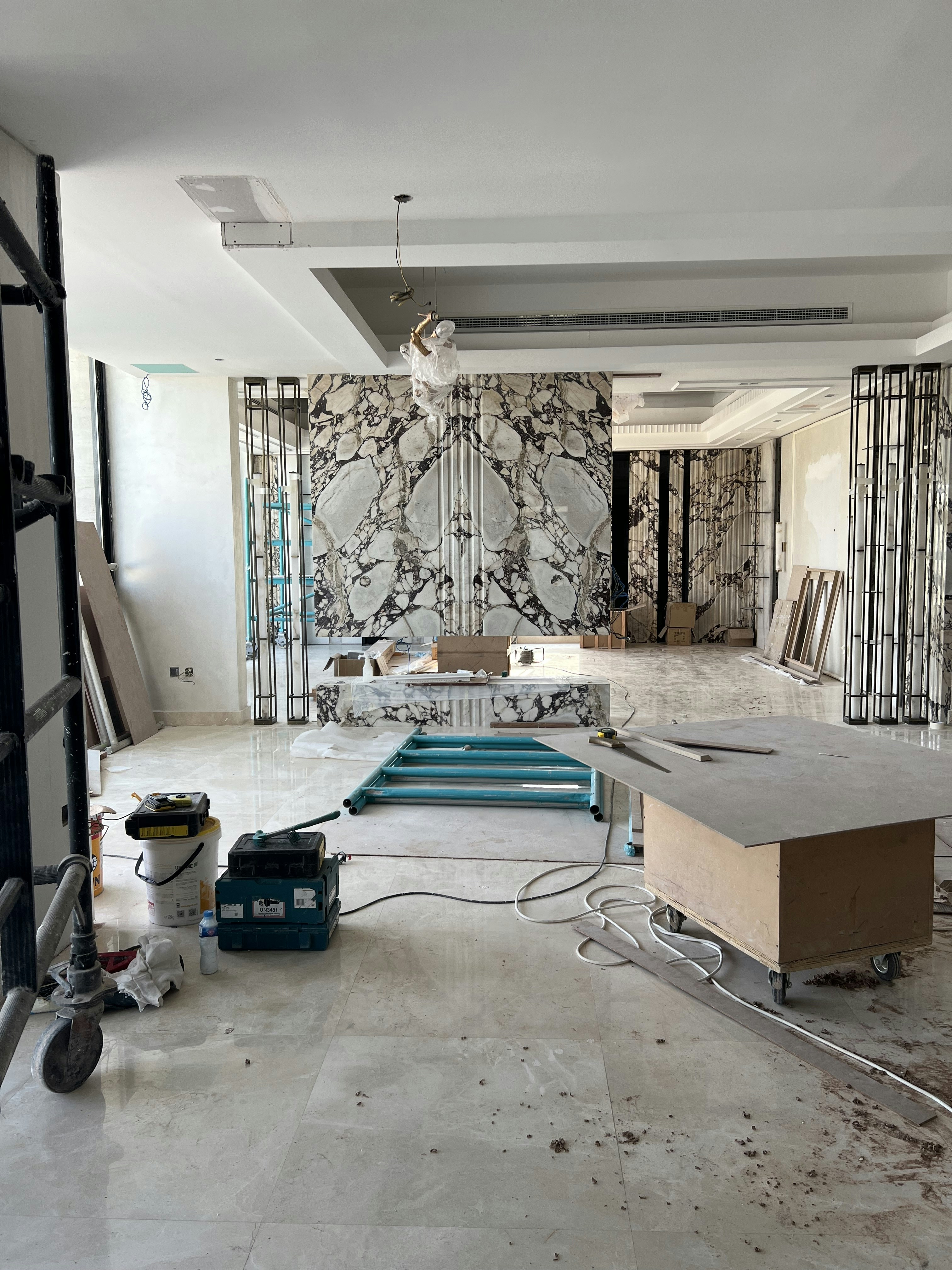 An interior space appears to be under renovation, featuring a large, decorative marble wall with an intricate pattern. Scaffolding, tools, and materials are scattered throughout the room, including a cart, power tools, and a table. The room is lit by natural light coming through large windows, adding to the sense of openness.