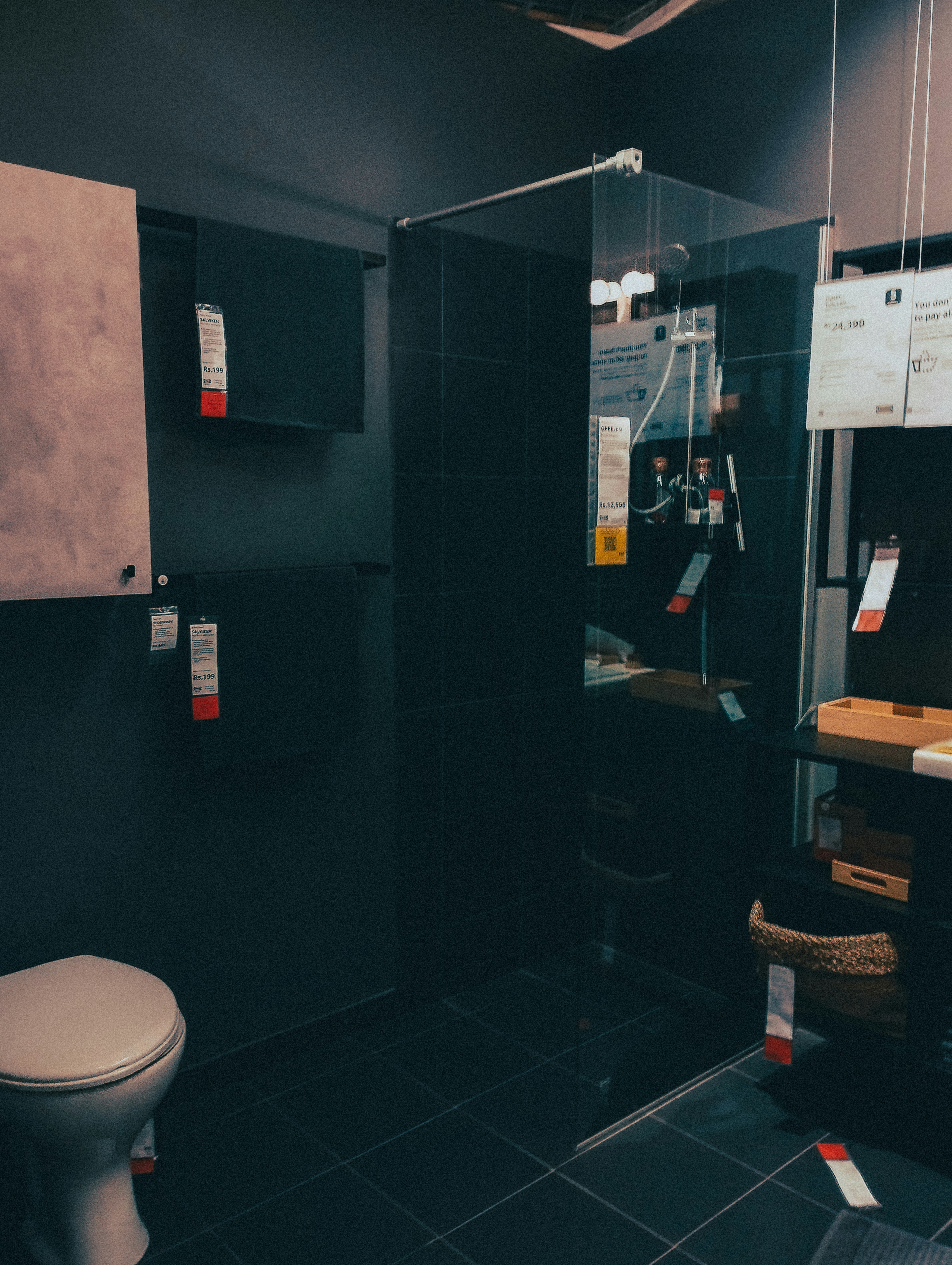 A modern bathroom showroom featuring a toilet, a glass shower enclosure, and dark tile flooring. The walls incorporate built-in cabinetry with minimalist designs. A wicker basket and some packaged products are visible under a counter.
