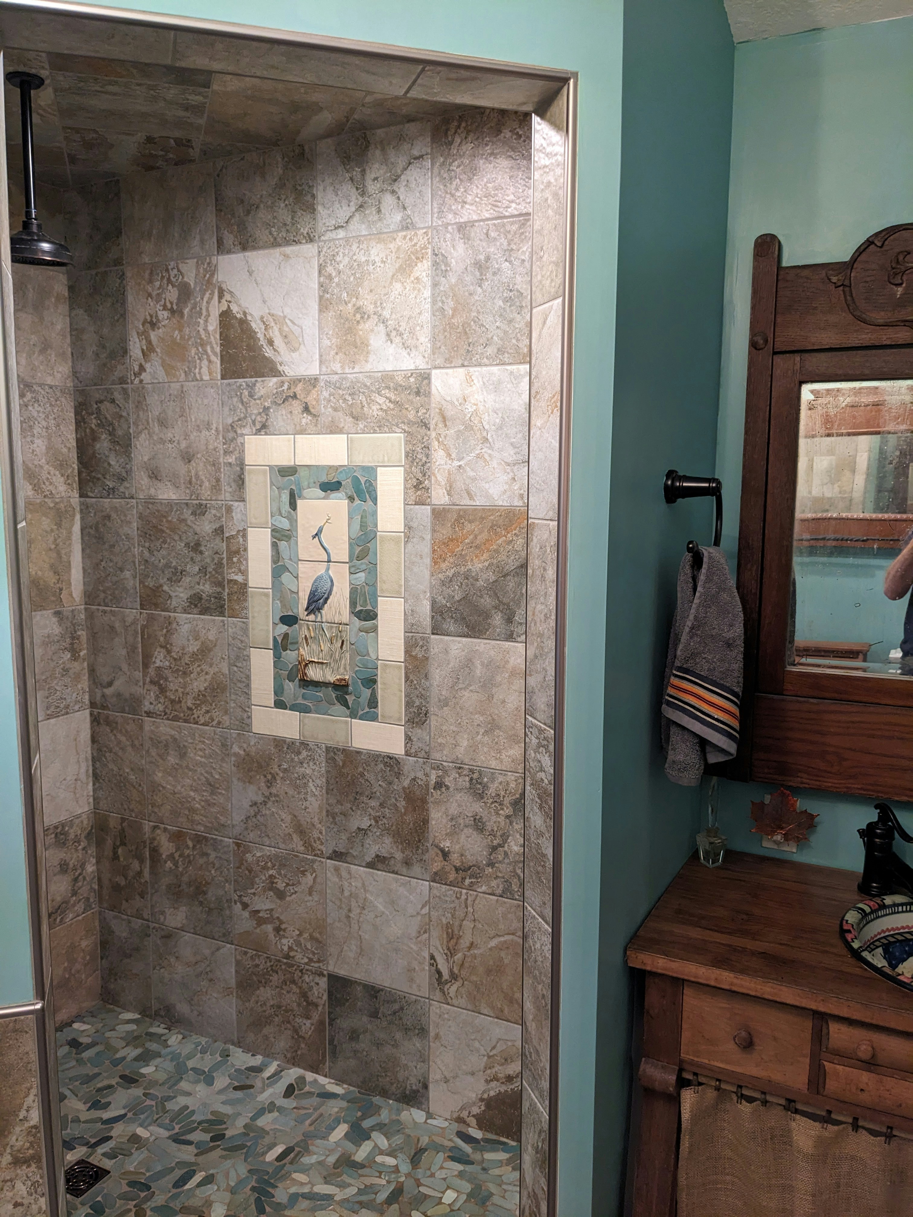 A bathroom featuring a tiled shower area with a decorative tile inlay depicting a bird in the center. The shower tiles are of neutral earth tones. To the right, there's a wood-framed mirror above a wooden cabinet with a towel hanging on a black holder. The room has walls painted in a soft teal color.